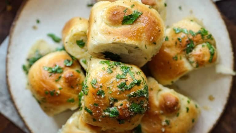 A plate of golden-brown garlic knots recipe topped with chopped parsley and melted butter, arranged on a round ceramic dish.
