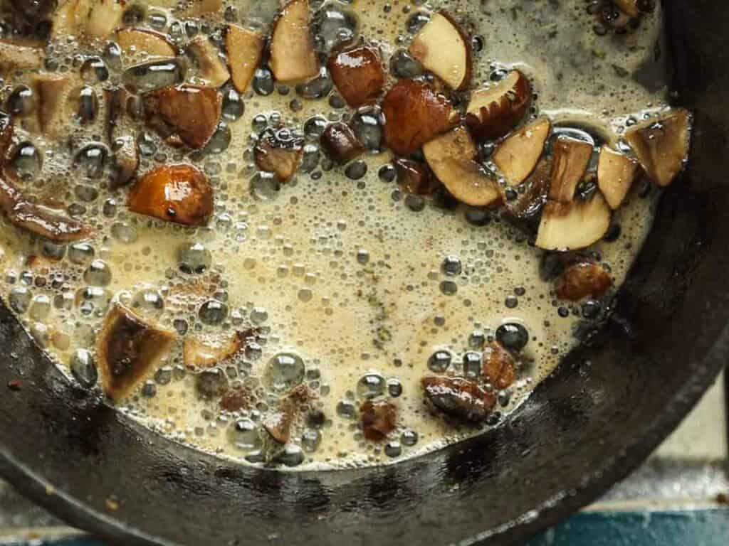 Chopped mushrooms cooking in a skillet with bubbling liquid, possibly butter or oil, creating a foamy texture around the pieces.