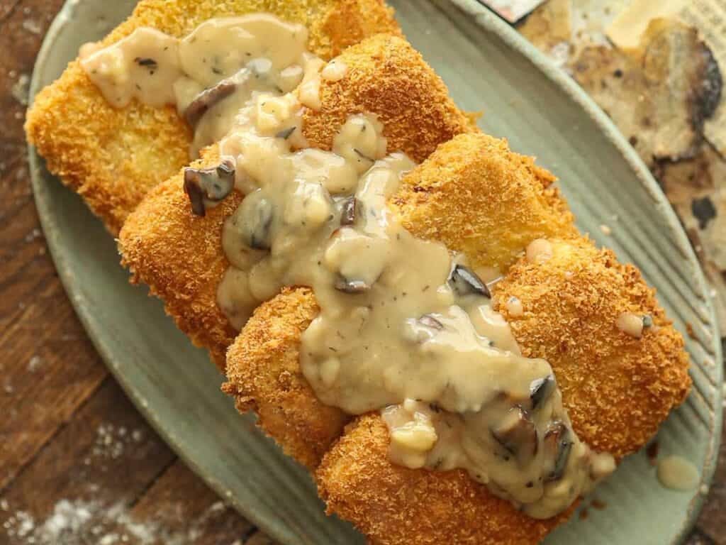 A breaded, golden-brown fried cutlet topped with creamy vegan mushroom sauce is served on an oval plate, set on a rustic wooden surface.