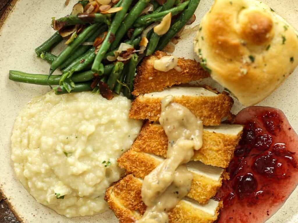 A plate with sliced breaded tof katsu topped with gravy, green beans with almond slivers, mashed potatoes, a dinner roll, and a serving of red berry sauce.