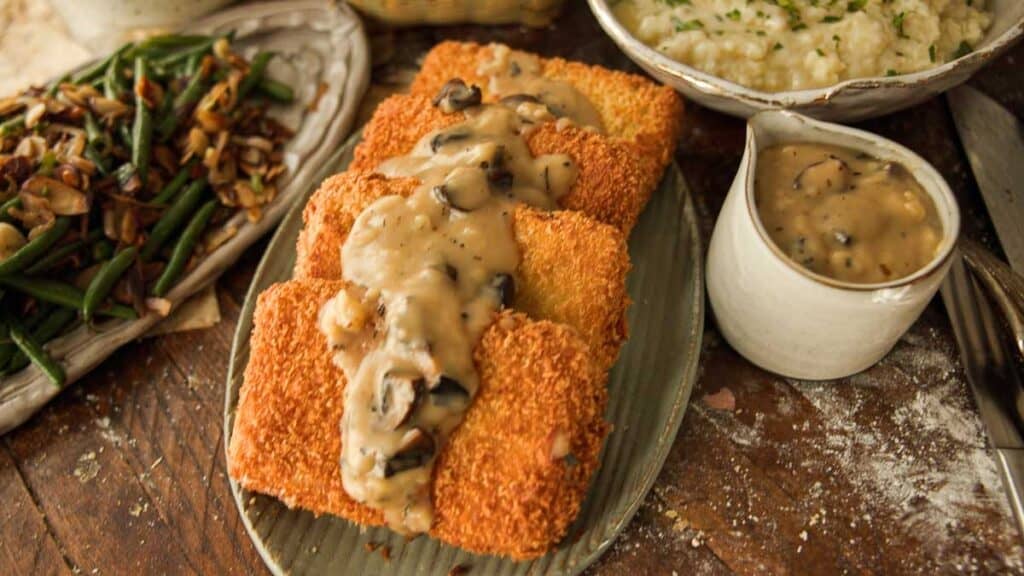 Three breaded and fried cutlets topped with creamy vegan mushroom gravy on an oval plate, with a side of green beans, mashed potatoes, and extra gravy in a small pitcher on a rustic wooden table.