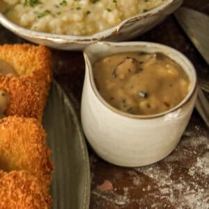 A small white ceramic pitcher filled with creamy vegan mushroom gravy sits on a rustic table next to breaded fried food and a bowl of mashed potatoes.