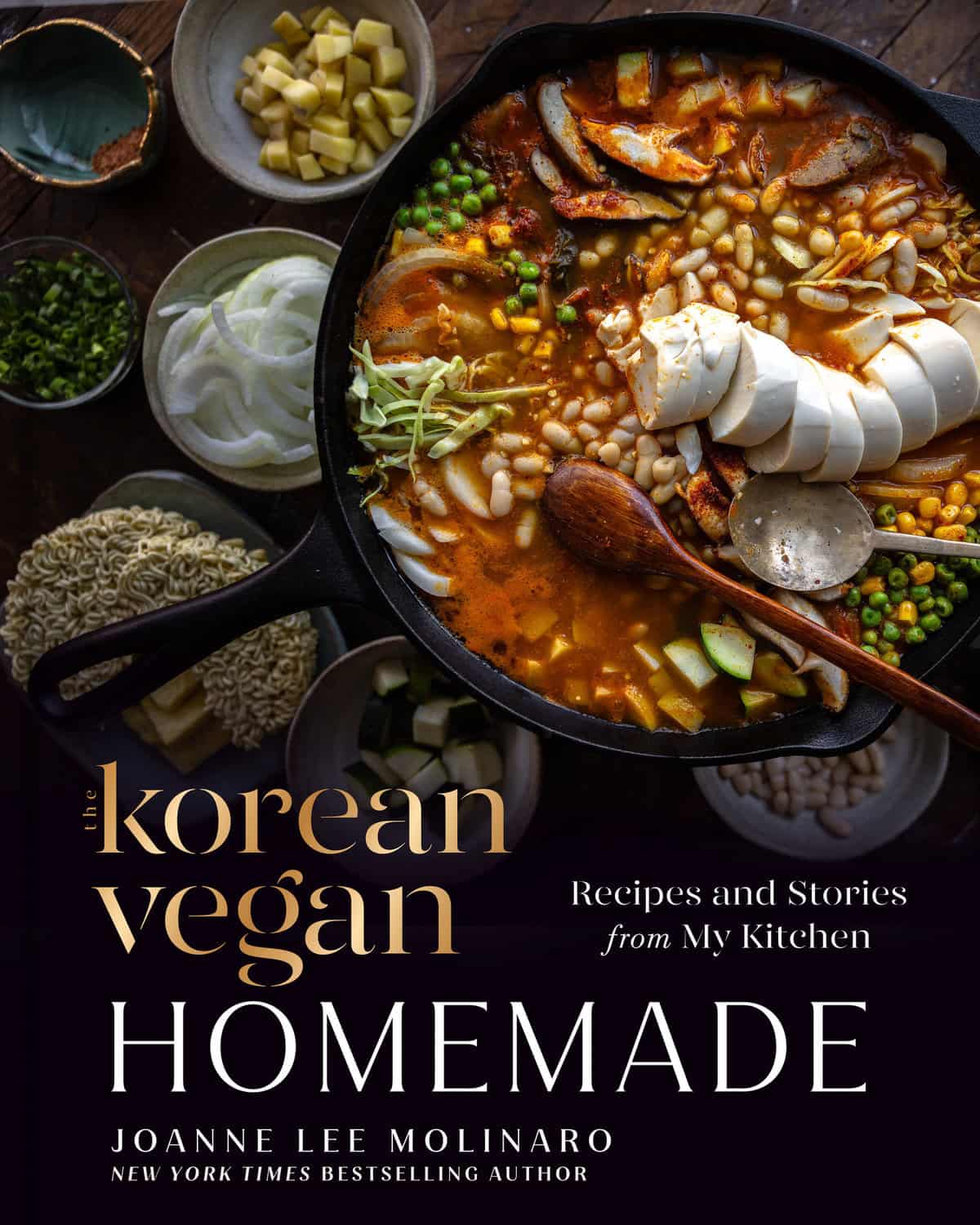 A skillet filled with a colorful Korean Vegan stew, topped with tofu, white beans, and mushrooms, surrounded by bowls of vegetables and noodles. Text reads: “The Korean Vegan: Homemade” by Joanne Lee Molinaro—perfect for any Vegan Holiday Gift!.