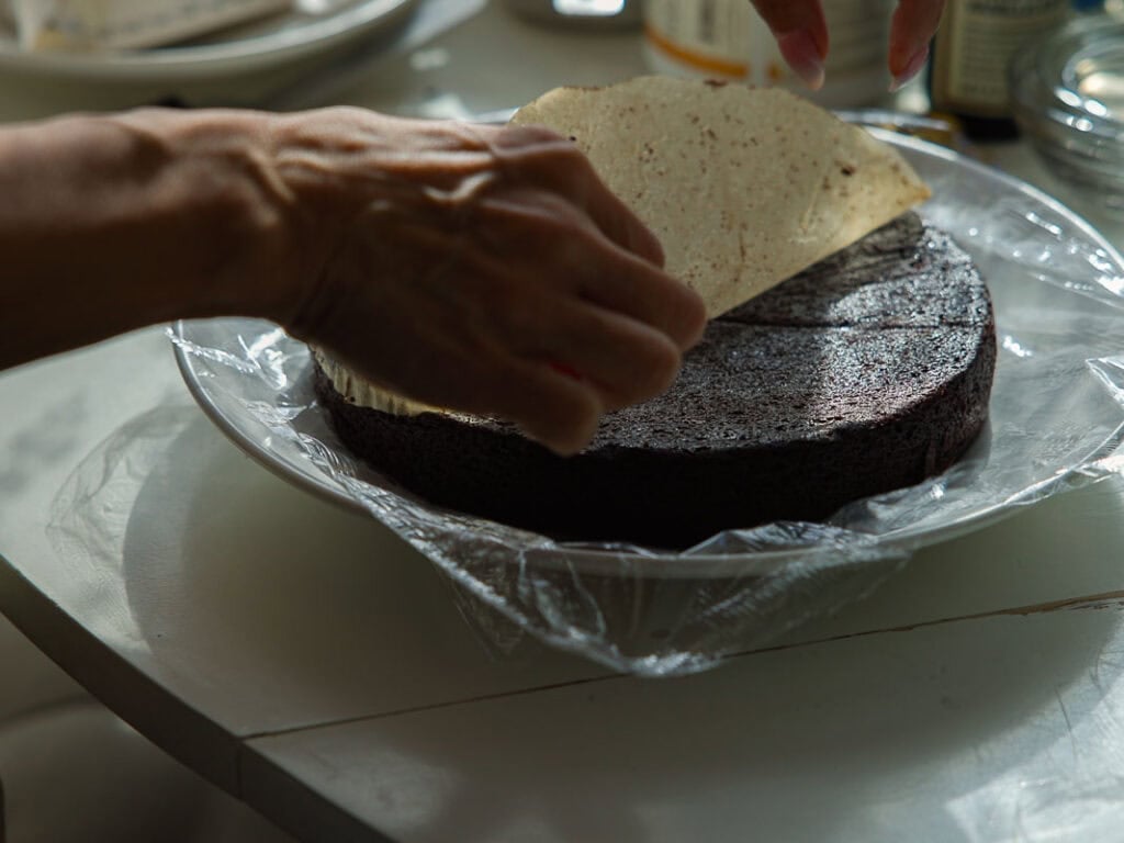 The BEST Vegan Chocolate Cake Recipe On The Internet. 23 A person’s hands are removing parchment paper circle on top of a round chocolate cake that sits on plastic wrap in a glass dish. The cake appears to be in the process of being assembled or decorated.