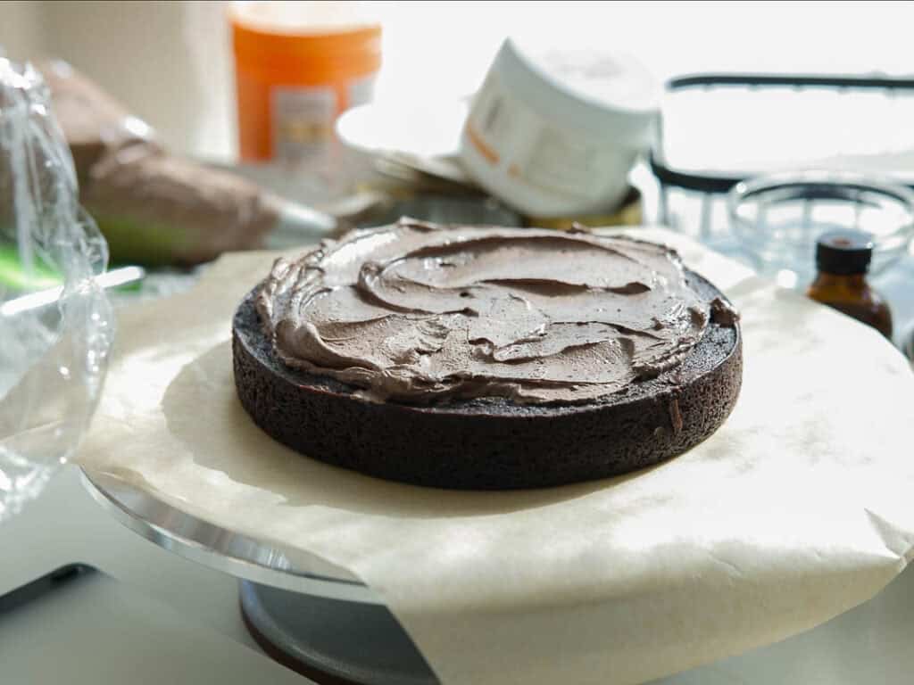 The BEST Vegan Chocolate Cake Recipe On The Internet. 30 A single round chocolate cake layer with chocolate frosting on top sits on parchment paper, surrounded by baking supplies in a bright kitchen.