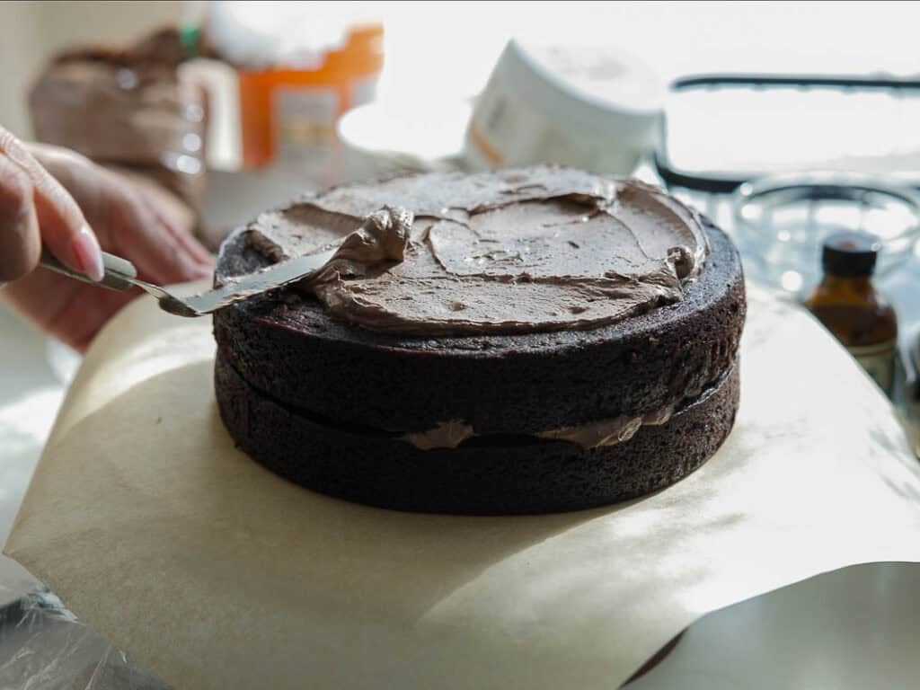 The BEST Vegan Chocolate Cake Recipe On The Internet. 31 A hand spreads chocolate frosting on a two-layer chocolate cake with a spatula. Baking supplies and ingredients are visible in the background on a sunlit kitchen counter.