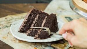 A hand holding a fork is about to take a bite from a slice of layered vegan chocolate cake on a white plate, set on top of old papers on a rustic wooden table.