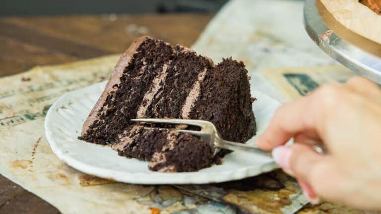 A hand holding a fork is about to take a bite from a slice of layered vegan chocolate cake on a white plate, set on top of old papers on a rustic wooden table.