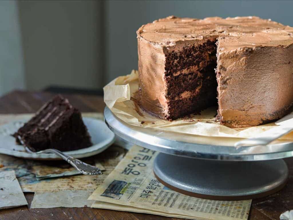The BEST Vegan Chocolate Cake Recipe On The Internet. 2 A large vegan chocolate layer cake with a slice missing sits on a cake stand. The missing slice is on a plate nearby, showing the cake’s rich, dark interior and chocolate frosting between layers. A fork rests beside the slice.