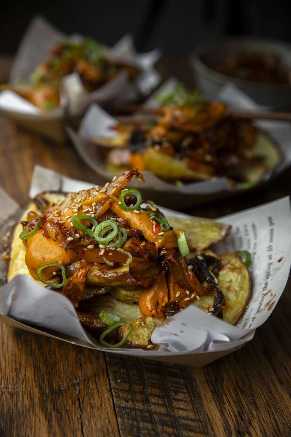 10+ Delicious and Easy Recipes Using Kimchi Today! 18 A close-up of crispy potato slices topped with shredded meat, sliced green onions, sesame seeds, and sauce—an easy recipe—served in a paper-lined tray on a rustic wooden table. Other similar trays are blurred in the background.