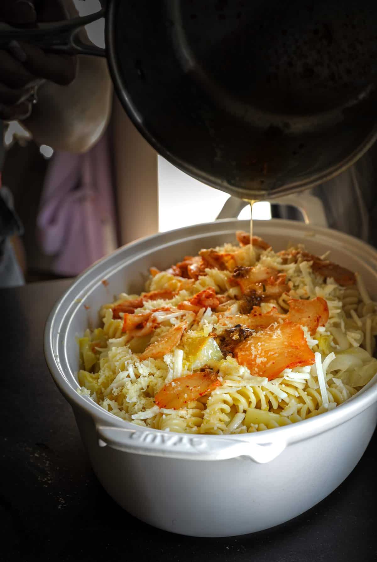 10+ Delicious and Easy Recipes Using Kimchi Today! 17 A hand pours sauce from a pan onto a white dish filled with rotini pasta, shredded cheese, and roasted vegetables—one of those easy recipes perfect for preparing a baked pasta dish.