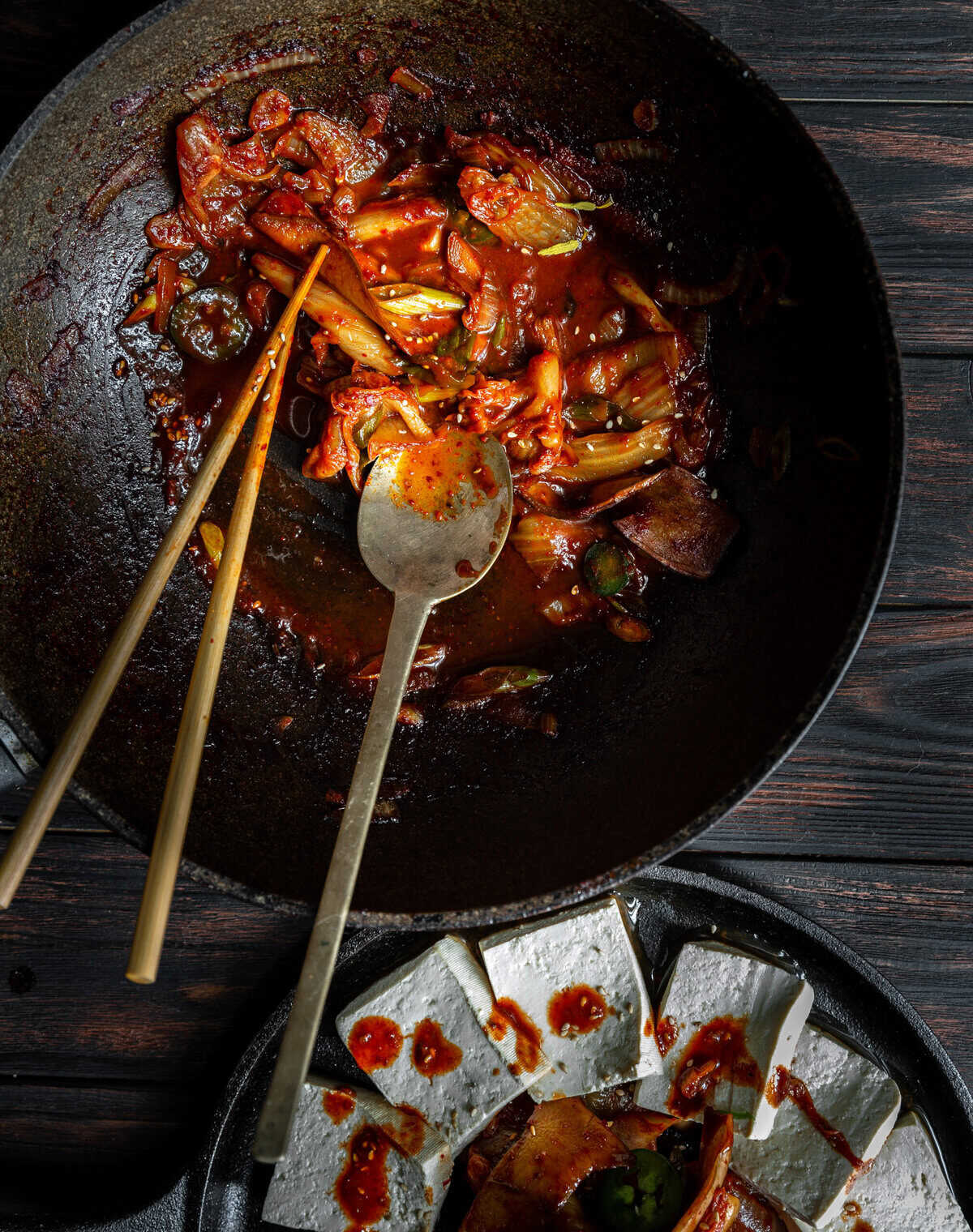 10+ Delicious and Easy Recipes Using Kimchi Today! 20 A wok with spicy stir-fried vegetables inspired by delicious kimchi, a metal spoon, and wooden chopsticks rests above a plate of sliced tofu drizzled with red sauce on a dark wooden table.