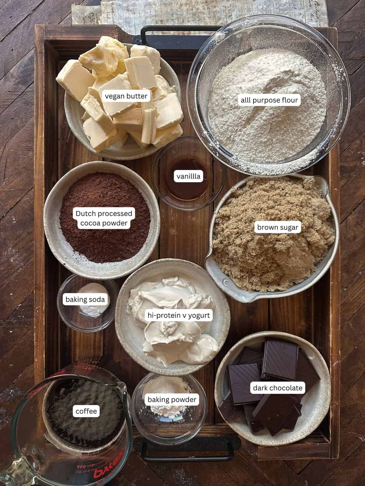 The BEST Vegan Chocolate Cake Recipe On The Internet. 4 A wooden tray holds bowls of ingredients for baking a vegan chocolate cake, including vegan butter, all-purpose flour, Dutch-processed cocoa powder, brown sugar, vanilla, baking soda, hi-protein yogurt, baking powder, coffee, and dark chocolate.