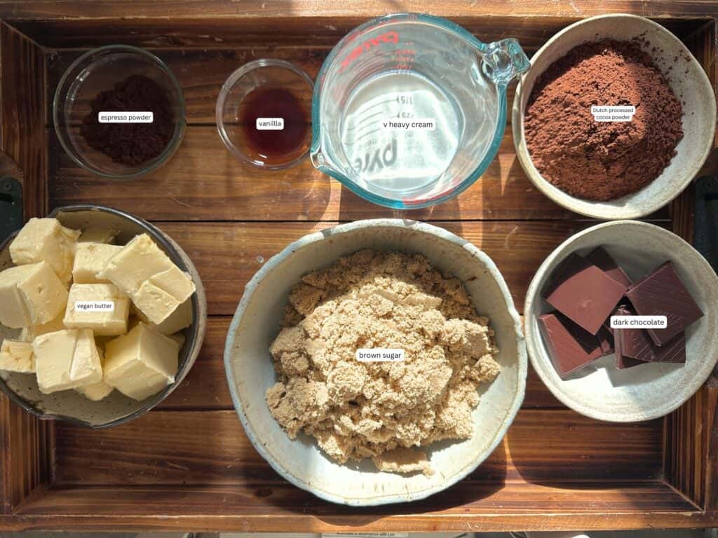 The BEST Vegan Chocolate Cake Recipe On The Internet. 5 A wooden tray with bowls and cups containing ingredients for vegan chocolate cake—vegan butter, brown sugar, cocoa powder, dark chocolate, heavy cream, vanilla extract, and espresso powder—each labeled with white stickers.
