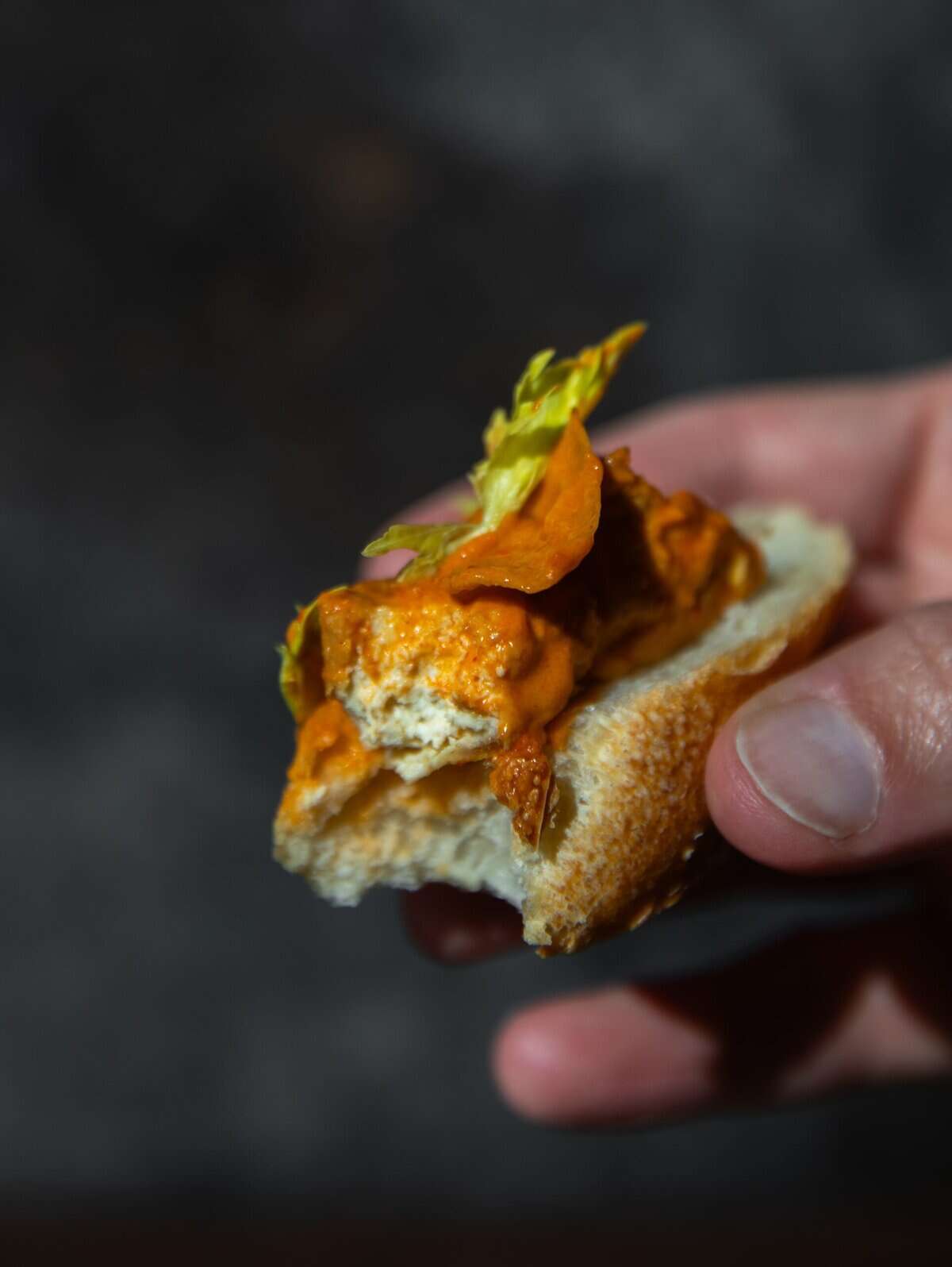 10+ Delicious and Easy Recipes Using Kimchi Today! 16 A hand holding a piece of bread topped with a creamy orange sauce inspired by delicious kimchi, herbs, and a leafy green garnish, against a dark, blurred background.