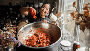 Joanne Lee Molinaro holds up a piece of kimchi over a large metal bowl filled with more kimchi, surrounded by jars, flowers, and kitchen items—perfect inspiration for new recipes using kimchi—on a table by a window.