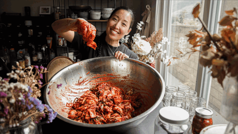 Joanne Lee Molinaro holds up a piece of kimchi over a large metal bowl filled with more kimchi, surrounded by jars, flowers, and kitchen items—perfect inspiration for new recipes using kimchi—on a table by a window.