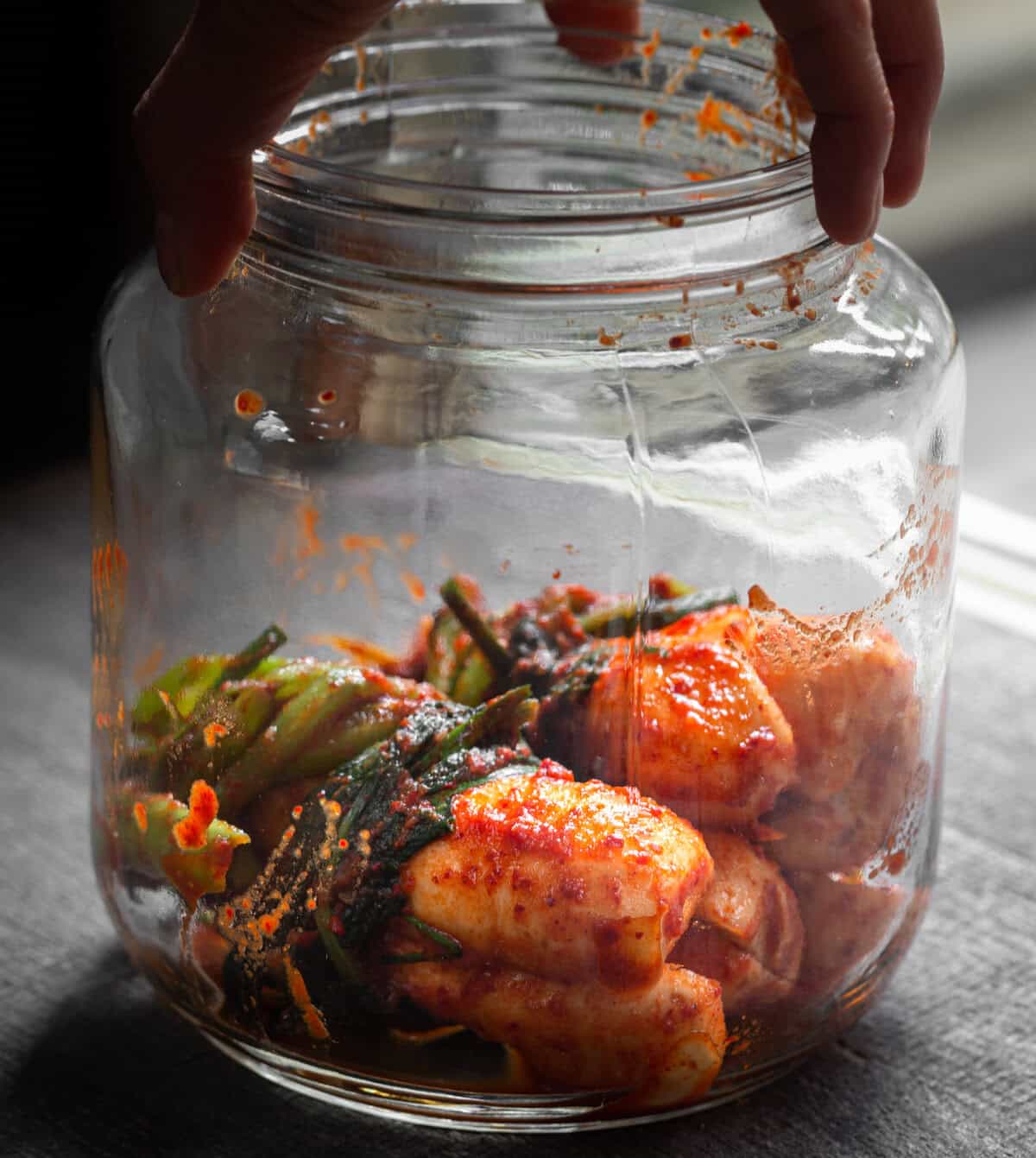 10+ Delicious and Easy Recipes Using Kimchi Today! 19 A hand holds a large glass jar of freshly made kimchi, with napa cabbage and green onions coated in spicy red seasoning. Light streams in, highlighting the vibrant colors—a perfect visual for delicious kimchi recipes.