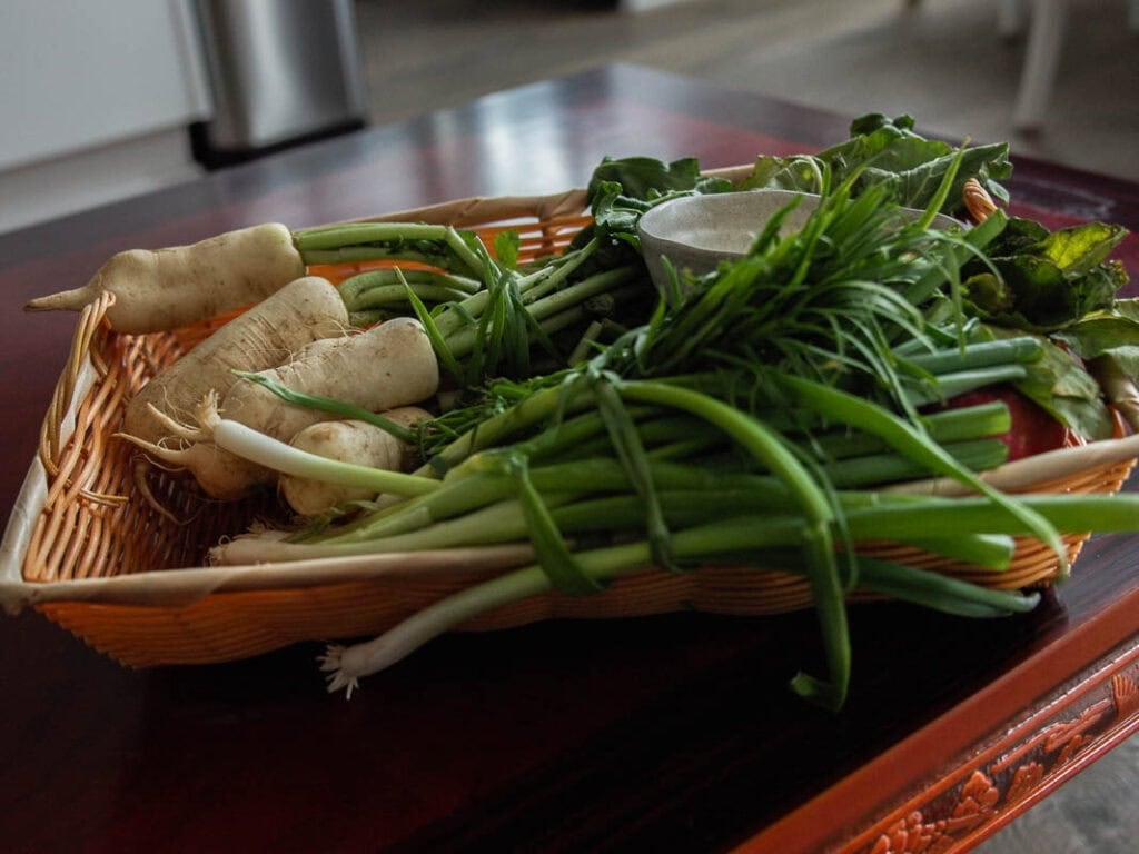 Vegan Ponytail Kimchi aka Chonggak-Kimchi Recipe. 4 A wicker basket on a table holds white radishes, green onions, leafy greens, and a small bowl—perfect for preparing Vegan Ponytail Kimchi in a cozy kitchen environment.