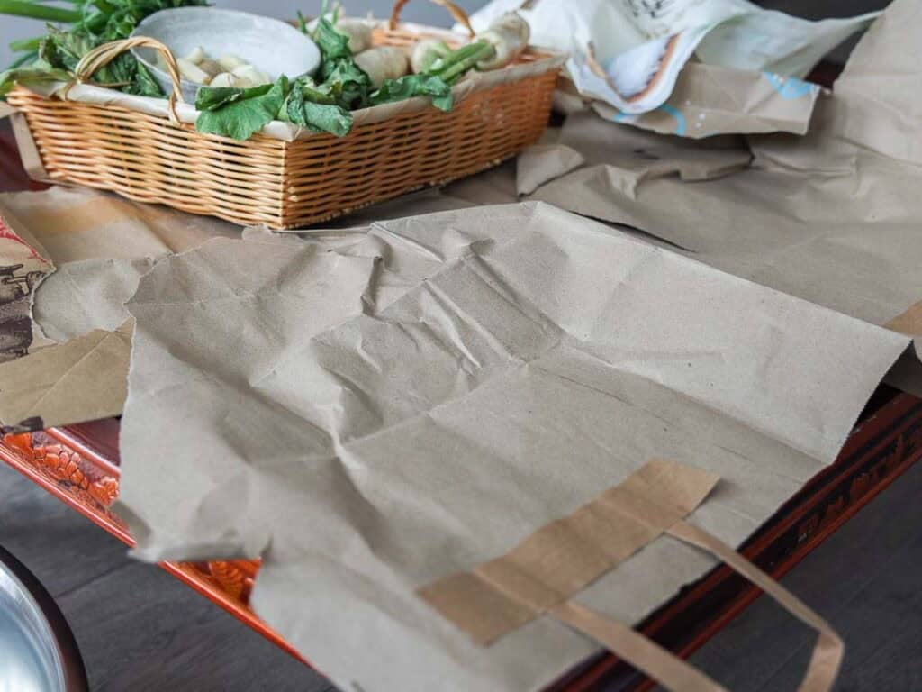 Vegan Ponytail Kimchi aka Chonggak-Kimchi Recipe. 13 Brown paper bags are spread out on a table next to a wicker basket filled with leafy vegetables and some dishes—perfect ingredients for making Vegan Ponytail Kimchi. Other paper and items are scattered on the surface.