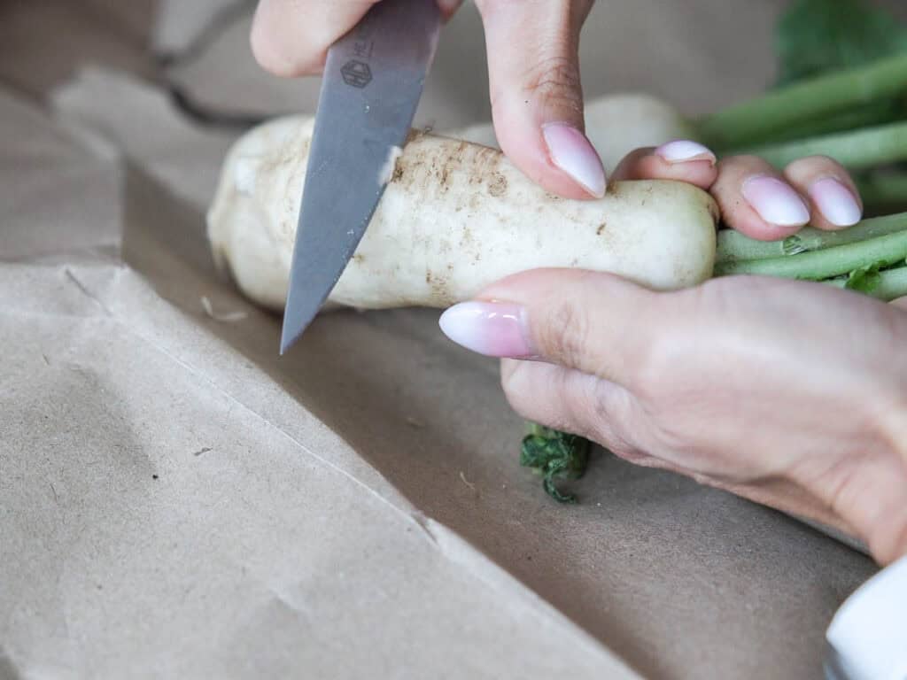Vegan Ponytail Kimchi aka Chonggak-Kimchi Recipe. 15 A person with light pink manicured nails slices a white radish with green stems on brown paper, prepping ingredients for a Vegan Kimchi recipe.