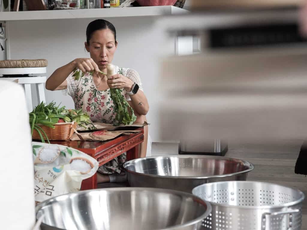 Vegan Ponytail Kimchi aka Chonggak-Kimchi Recipe. 17 A woman in an apron with a neat ponytail sits on the floor, peeling green vegetables for her Vegan Ponytail Kimchi. Metal bowls, fresh produce, and a bag of rice with Korean writing surround her in this cozy indoor kitchen setting.