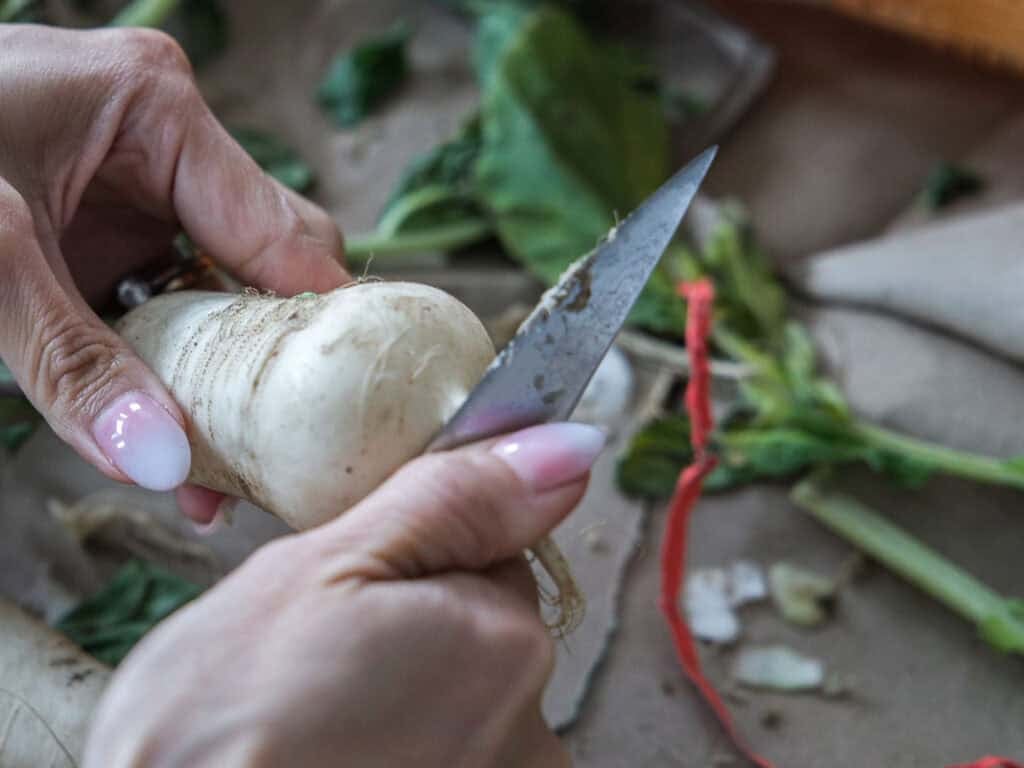 Vegan Ponytail Kimchi aka Chonggak-Kimchi Recipe. 14 Close-up of hands peeling a white root vegetable with a knife, surrounded by leafy greens and scraps—perfect preparation for a Vegan Kimchi or trying out an authentic Chonggak-Kimchi Recipe.