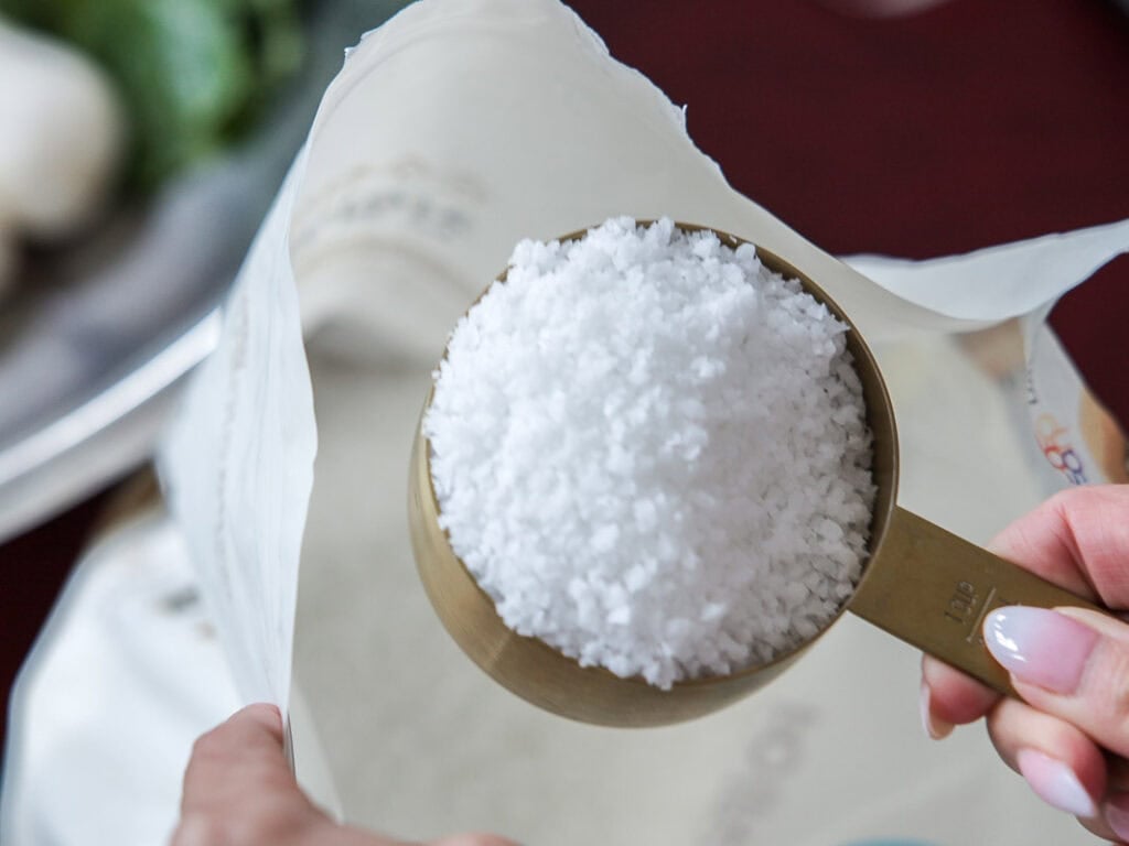 Vegan Ponytail Kimchi aka Chonggak-Kimchi Recipe. 6 A hand holding a gold measuring cup filled with coarse sea salt over an open bag, with blurred green vegetables in the background—perfect for preparing Vegan Ponytail Kimchi using an authentic Chonggak-Kimchi recipe.