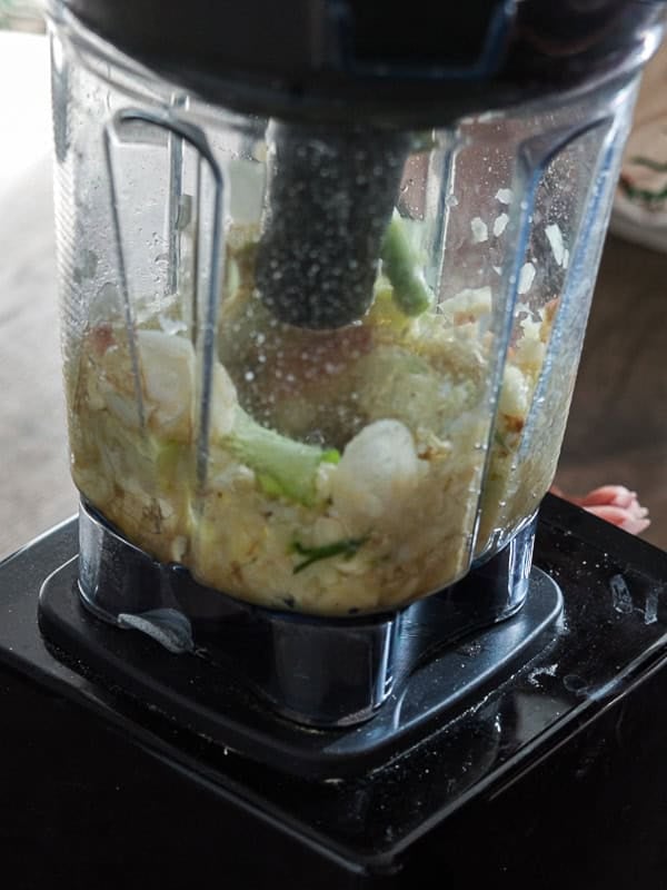Vegan Ponytail Kimchi aka Chonggak-Kimchi Recipe. 26 Close-up of a blender mixing chopped vegetables, including green onions and garlic, on a black base. The ingredients are partially blended for a Vegan Kimchi, with condensation and food splatters visible on the pitcher.