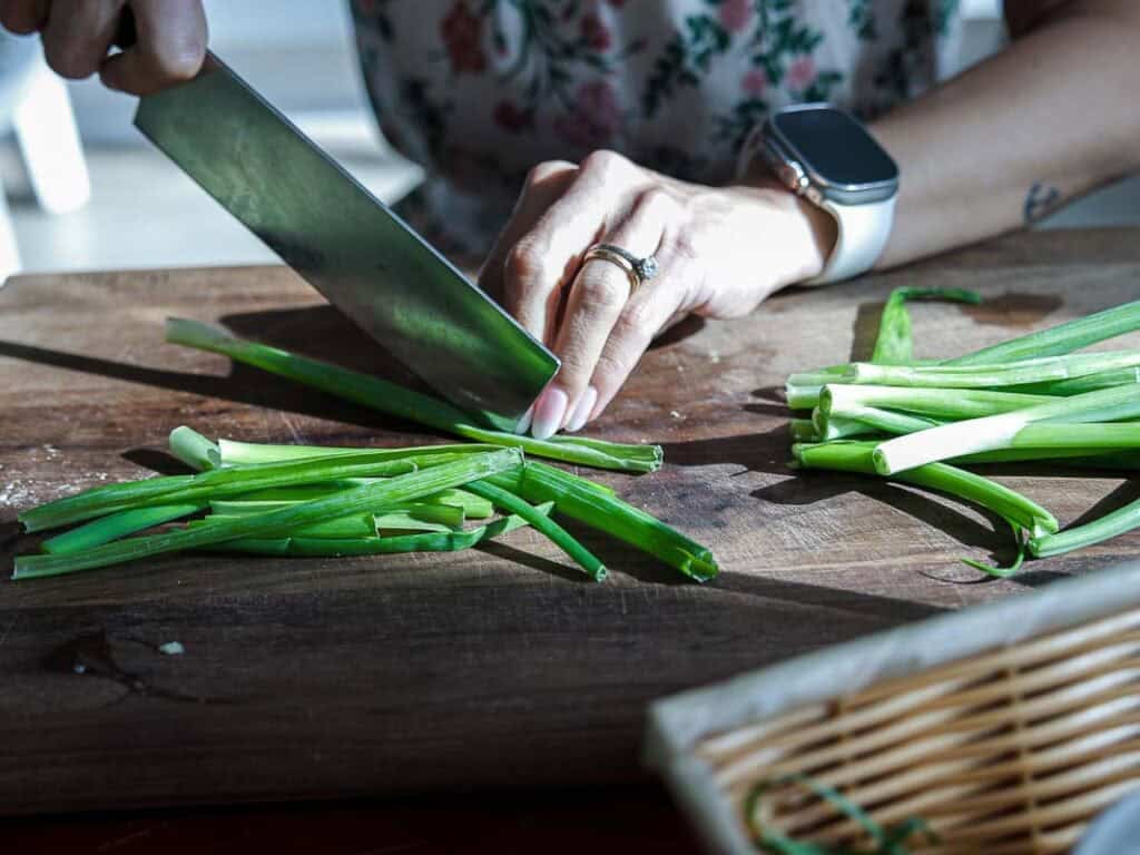 Vegan Ponytail Kimchi aka Chonggak-Kimchi Recipe. 7 A person wearing a ring and smartwatch is slicing green onions on a wooden cutting board, preparing ingredients for a Vegan Kimchi. Some chopped green onions are gathered on the side, and a woven basket is partially visible in the foreground.