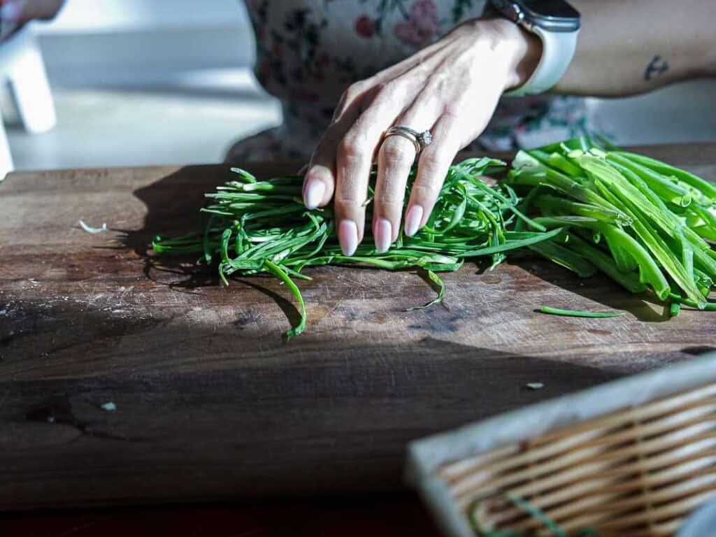 Vegan Ponytail Kimchi aka Chonggak-Kimchi Recipe. 10 A person wearing a ring and a smartwatch arranges chopped green vegetables on a wooden cutting board, likely preparing ingredients for a vegan kimchi recipe, with a wicker basket partially visible in the foreground.