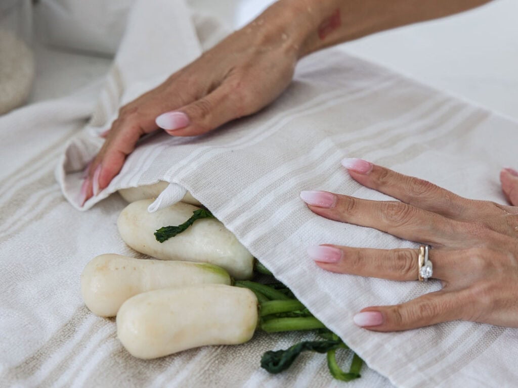 Vegan Ponytail Kimchi aka Chonggak-Kimchi Recipe. 34 A person with manicured nails and a ring wraps white radishes with green stems in a striped kitchen towel on a white surface, preparing ingredients for an authentic chonggak kimchi recipe.