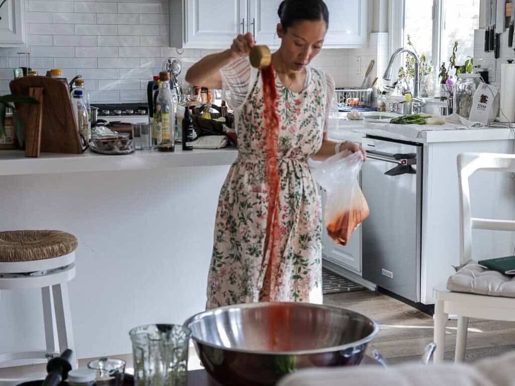 Vegan Ponytail Kimchi aka Chonggak-Kimchi Recipe. 8 A woman in a floral dress stands in a bright kitchen, pouring gochugaru from a measuring cup into a large metal bowl, preparing her vegan kimchi recipe. She holds a plastic bag with more red liquid, surrounded by various utensils.