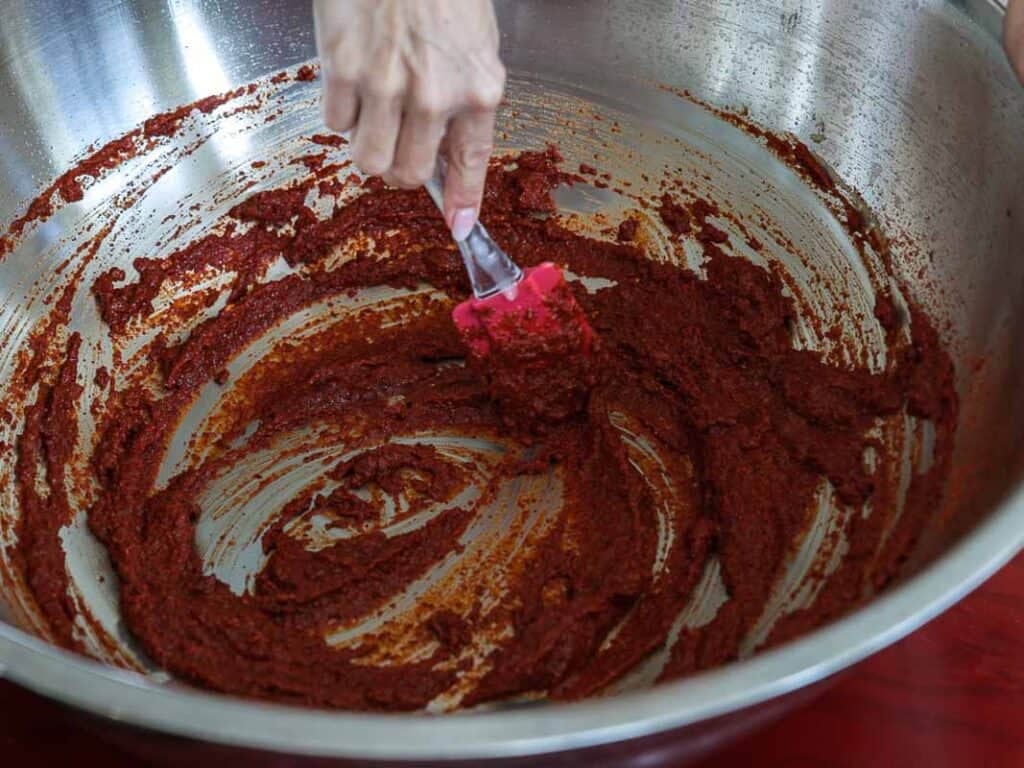 Vegan Ponytail Kimchi aka Chonggak-Kimchi Recipe. 39 A hand uses a red spatula to mix a thick, reddish-brown paste inside a large metal bowl while preparing Vegan Kimchi.