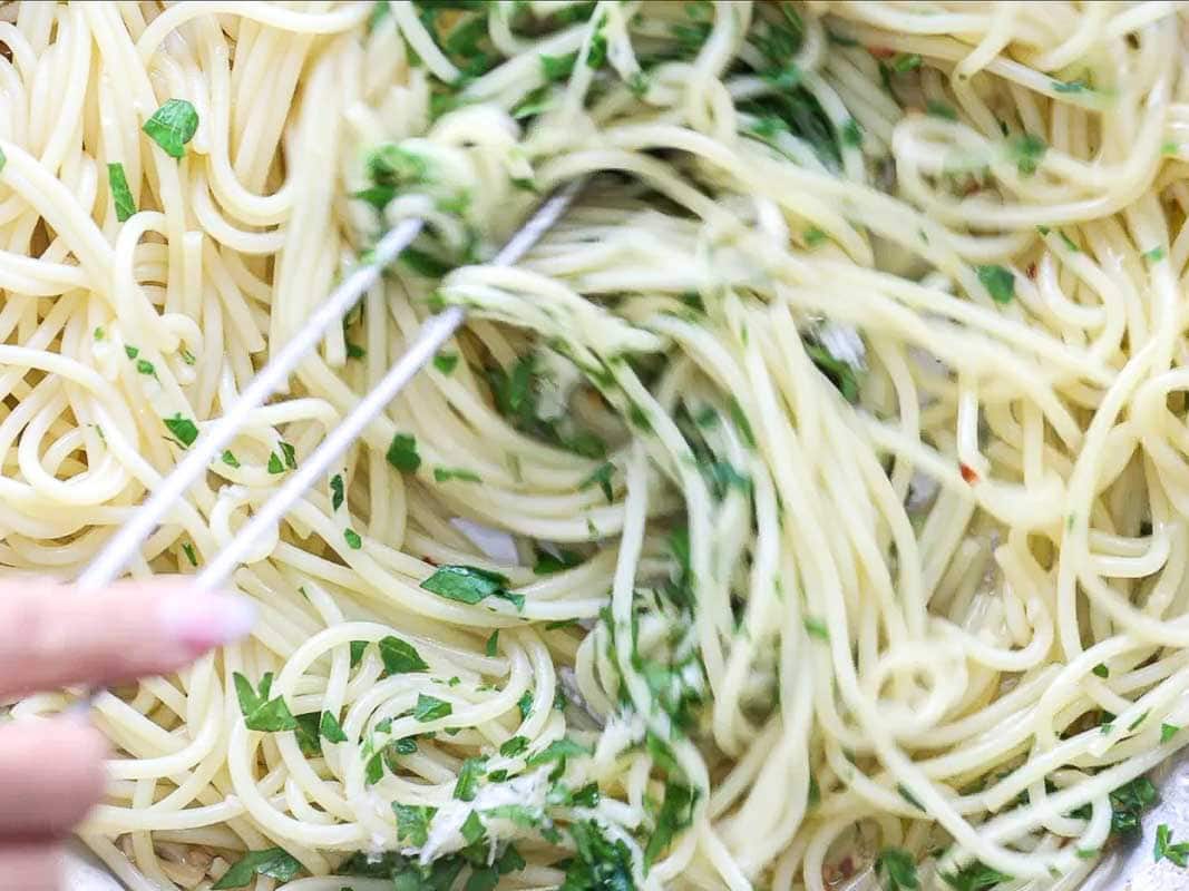 8-ingredient Easy and Delicious Lemon Pepper Pasta. 15 A close-up of delicious pasta being tossed with fresh chopped parsley using a fork and spoon. The glossy noodles are lightly coated, with visible green herbs mixed throughout for an easy pasta dish.