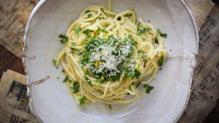 A bowl of easy pasta topped with grated cheese, chopped parsley, and lemon zest, served in a rustic ceramic dish on a table with old paper sheets underneath.