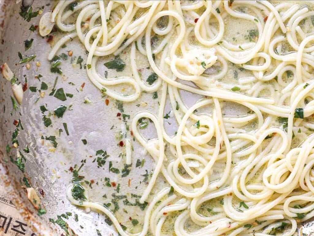 8-ingredient Easy and Delicious Lemon Pepper Pasta. 3 Cooked spaghetti noodles in a creamy, herb-filled sauce are scattered in a metal pan, with some sauce remnants visible and parts of the pan partially scraped clean—an easy pasta perfect for any night.