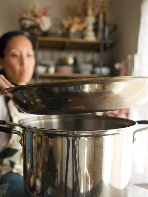 8-ingredient Easy and Delicious Lemon Pepper Pasta. 5 A woman lifts a large metal pan over a stainless steel pot, preparing Easy Pasta in a kitchen with shelves of dishes and plants in the background. The focus is on the pot and pan, with the woman slightly blurred.