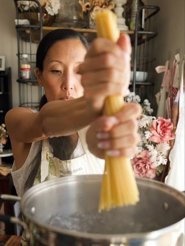 8-ingredient Easy and Delicious Lemon Pepper Pasta. 9 A woman in an apron holds a bundle of uncooked spaghetti above a pot of boiling water, ready to make an easy pasta recipe in a kitchen decorated with flowers and plants.