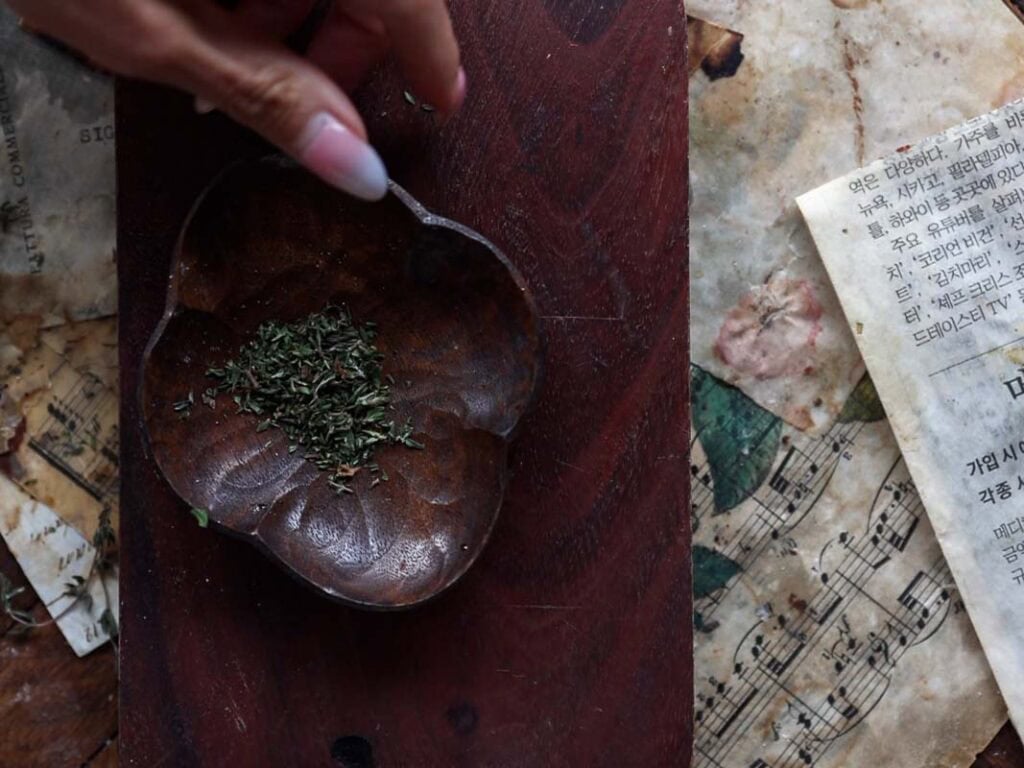 Easy Vegan Lemon Loaf Recipe - Better Than Starbucks 5 A hand sprinkles dried herbs into a carved wooden bowl on a dark wooden surface, with vintage sheet music, weathered papers written in Korean, and a vegan lemon loaf nearby.