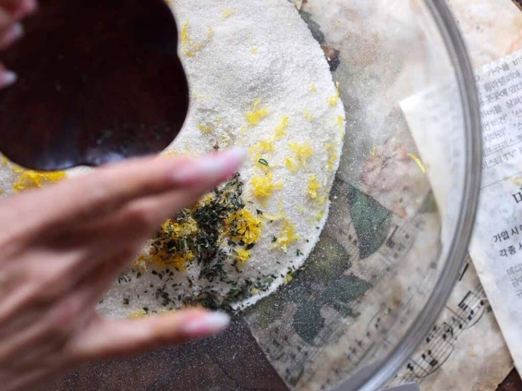 Easy Vegan Lemon Loaf Recipe - Better Than Starbucks 7 A hand sprinkles herbs into a glass bowl filled with sugar, lemon zest, and dried herbs—the perfect mix for a vegan lemon loaf. Pieces of newspaper and sheet music sit underneath the bowl.
