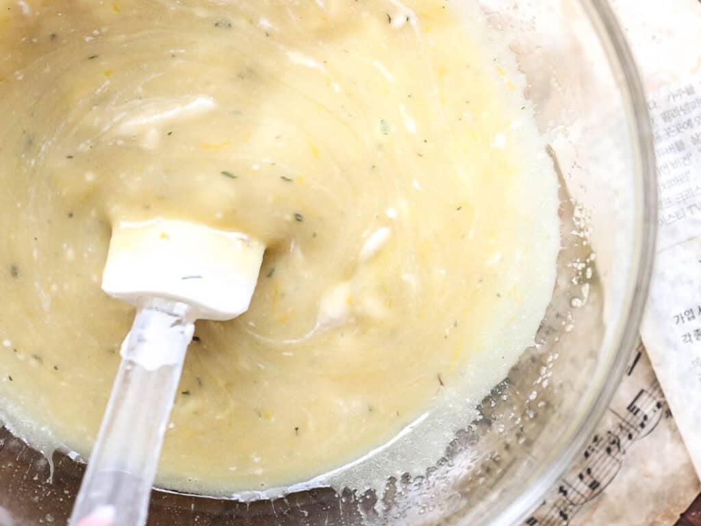 Easy Vegan Lemon Loaf Recipe - Better Than Starbucks 11 A glass bowl containing a thick, pale yellow vegan lemon loaf batter speckled with herbs, being mixed with a white spatula. Some batter clings to the sides of the bowl.