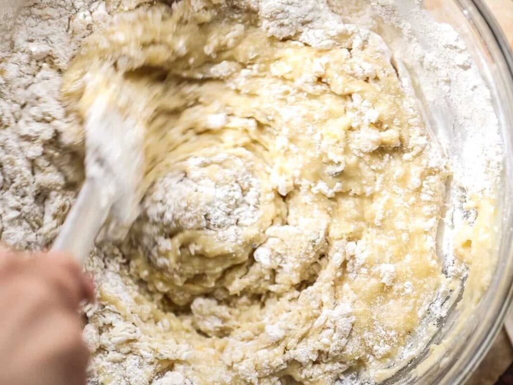 Easy Vegan Lemon Loaf Recipe - Better Than Starbucks 15 A hand stirs a thick, lumpy batter in a glass bowl, mixing flour into a yellowish mixture for a vegan lemon loaf with a spatula.