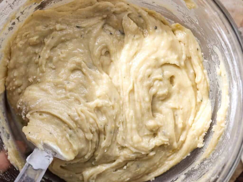 Easy Vegan Lemon Loaf Recipe - Better Than Starbucks 16 A glass bowl filled with thick, creamy batter for a vegan lemon loaf is being mixed with a spatula.