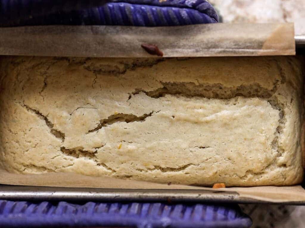 Easy Vegan Lemon Loaf Recipe - Better Than Starbucks 18 A close-up of a vegan lemon loaf with a cracked golden-brown crust, sitting in a parchment-lined baking pan. The pan is being held with a blue oven mitt.
