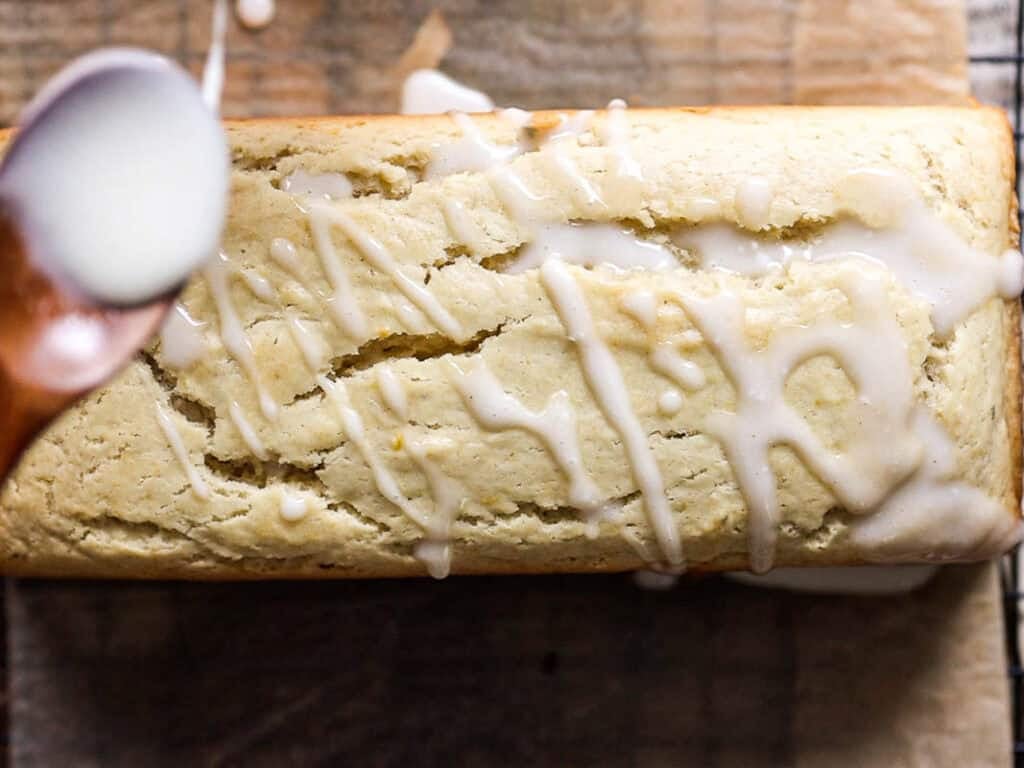 Easy Vegan Lemon Loaf Recipe - Better Than Starbucks 23 A vegan lemon loaf on parchment paper is being drizzled with white icing from a spoon, partially covering the top of the cake.