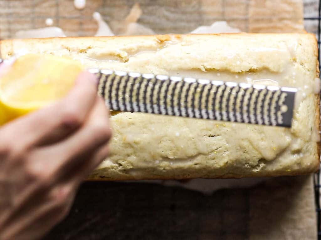 Easy Vegan Lemon Loaf Recipe - Better Than Starbucks 24 A hand holds a lemon and a zester over a glazed vegan lemon loaf on a wooden surface, grating fresh lemon zest onto the cake.