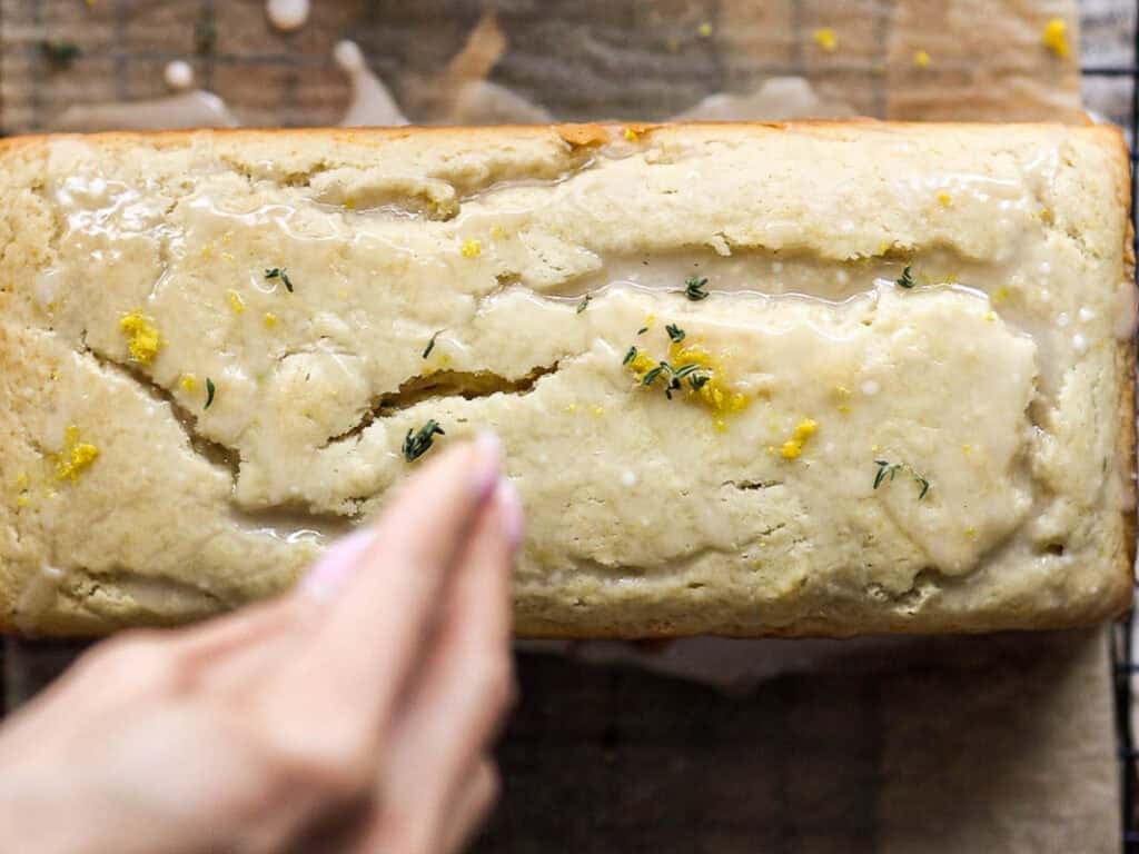 Easy Vegan Lemon Loaf Recipe - Better Than Starbucks 25 A hand sprinkles fresh thyme over a vegan lemon loaf glazed and topped with lemon zest, sitting on parchment paper.
