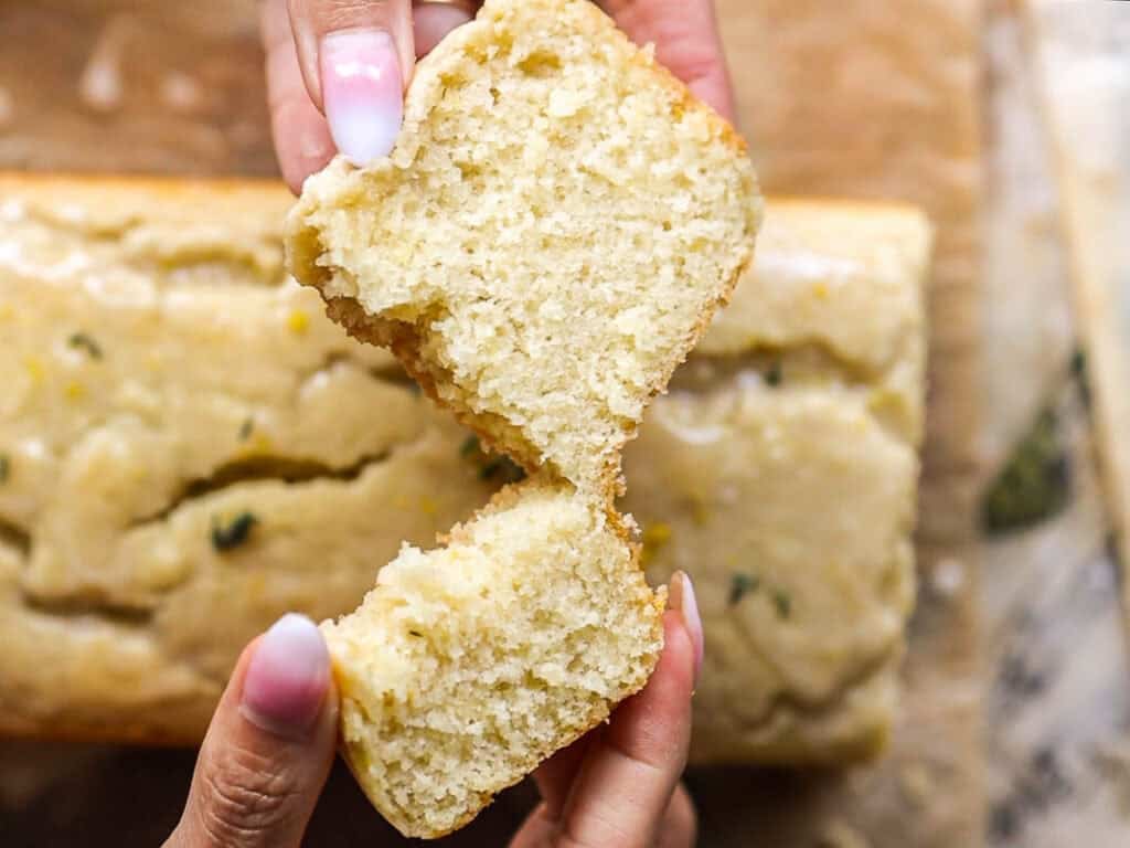 Easy Vegan Lemon Loaf Recipe - Better Than Starbucks 26 Two hands are pulling apart a moist, fluffy slice of vegan lemon loaf, revealing its soft, crumbly inside. A loaf with a smooth, glazed top sits in the background on a wooden surface.