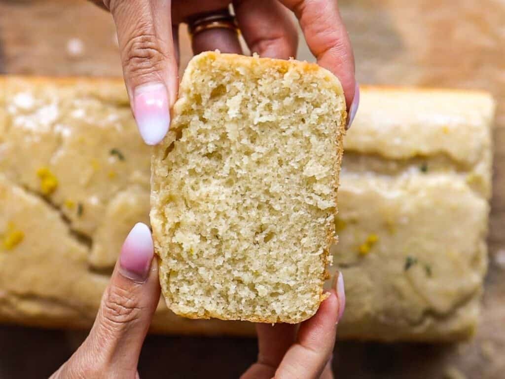 Easy Vegan Lemon Loaf Recipe - Better Than Starbucks 6 A person holding a thick slice of moist, golden vegan lemon loaf with light icing on top; the rest of the loaf is visible in the background on a wooden surface.